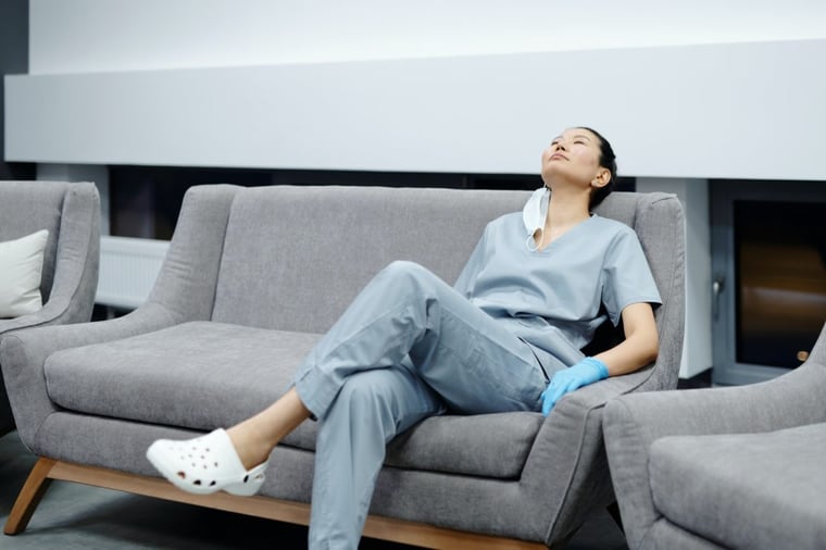 Tired nurse wearing gloves scrubs and mask rests her head on waiting room sofa