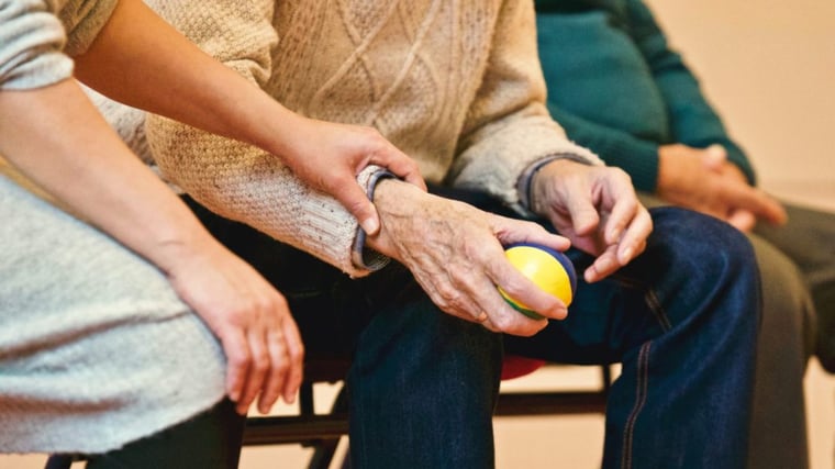 Elderly patient sits next to healthcare worker while she holds his hand as he holds a stress ball during physical therapy